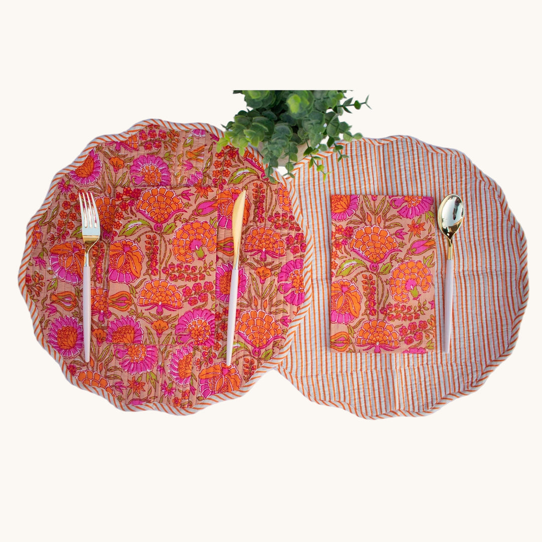 Reversible round quilted placemats with bold orange and magenta floral print on one side and orange stripes on the other, styled with cutlery and a plant.
