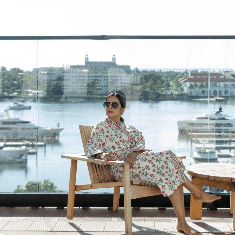 Woman in a floral robe sitting on a chair by a waterfront with buildings and water in the background.