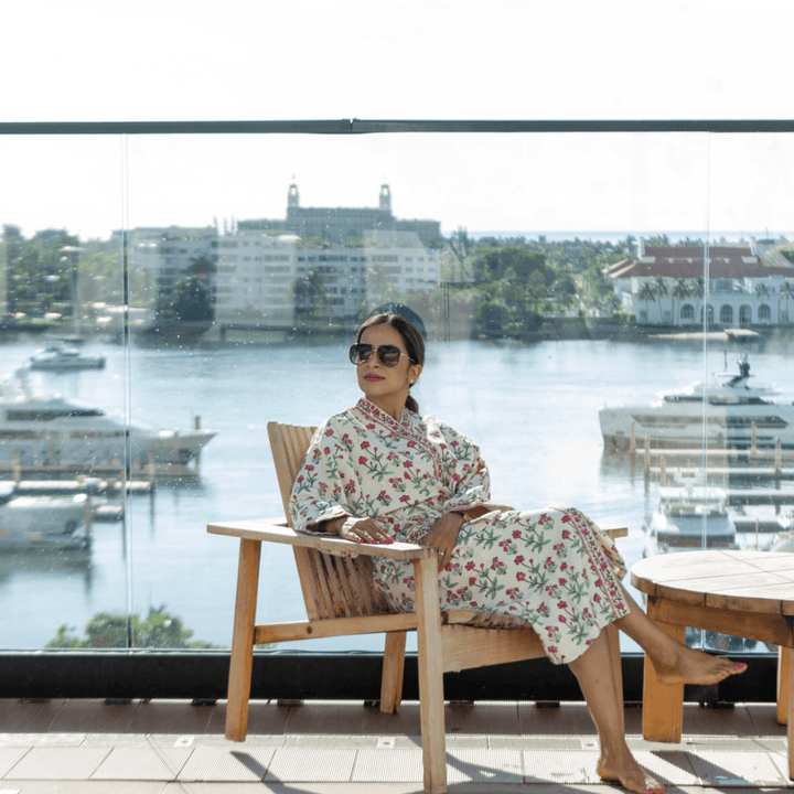 Woman in a floral robe sitting on a chair by a waterfront with buildings and water in the background.