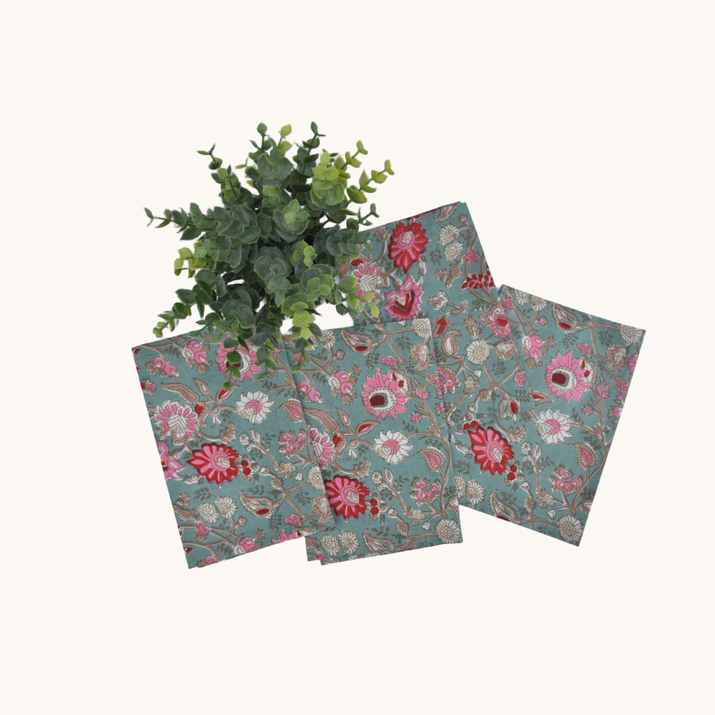 Set of four cotton napkins with teal background and pink floral block print, styled with a green potted plant
