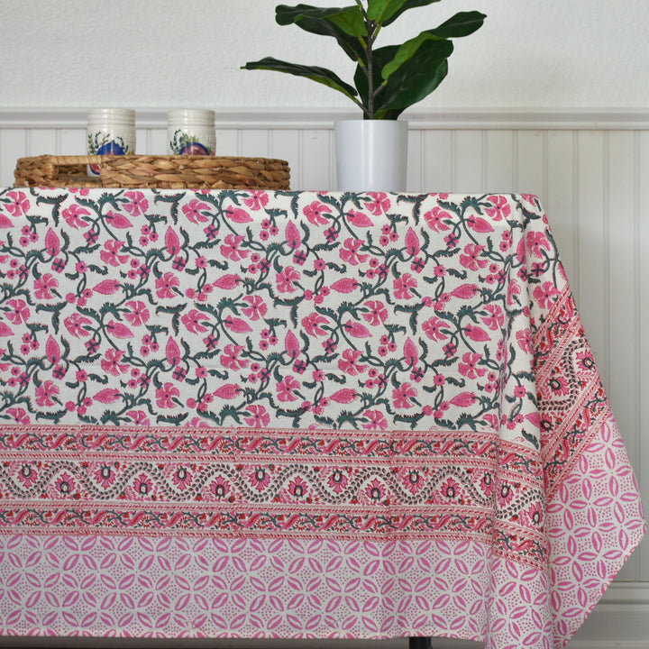Blossom Trail handblock print cotton tablecloth with pink floral pattern and intricate border, displayed on dining table with plant and basket decor.
